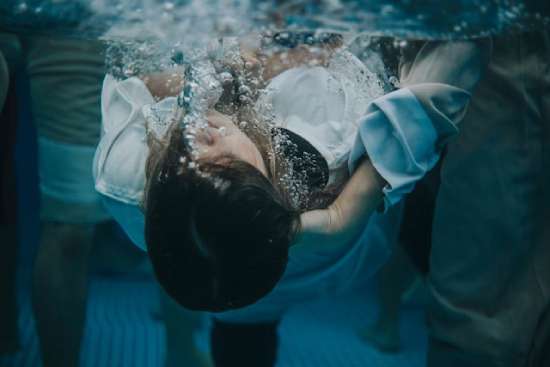 A young woman in a white robe is submerged underwater by a hand, signifying baptism's idea of death and burial.