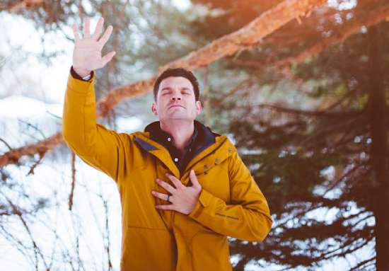 A man stands in a forest with his eyes closed and right hand raised up to show he's praying.