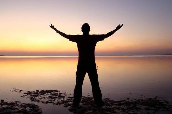 A silhouette of a man standing in front of a sunset over a lake with his arms reaching up. This is a common image to illustrate spiritual health.