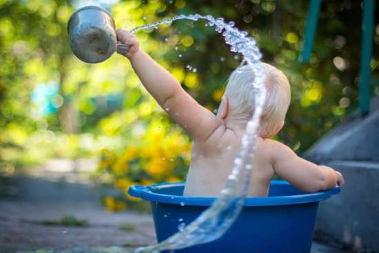 A baby in a small tub uses a cup to throw water.