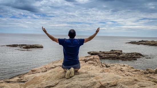  A man kneeling on a rocky shore, hands raised to the sky in repentance.