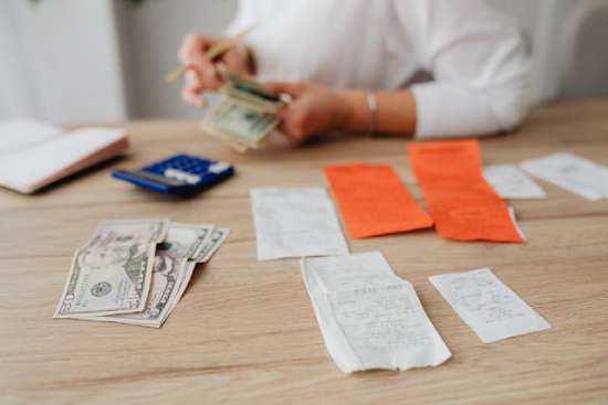 A person counts bills and organizes them on a tabletop. Stacks of receipts, a calculator, and a checkbook are also present.