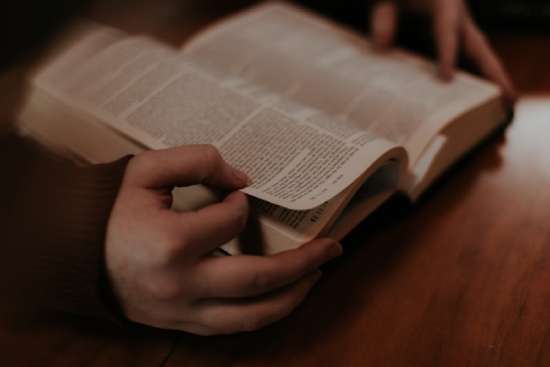 A close-up of hands holding an open Bible. Thumb and index finger are prepared to flip the page to continue studying the Bible.