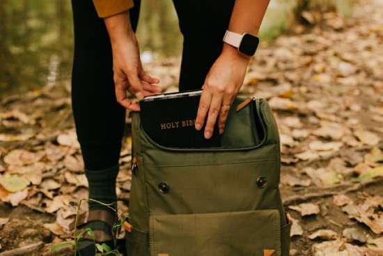 A green backpack sits at someone's feet. The person is leaning over to pull a Bible out of the backpack, highlighting the importance of searching the Word of God for answers, not the world.