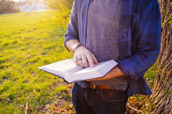 A person stands outside, holding an open Bible. Their finger is pointing to a verse that they seem to be considering.