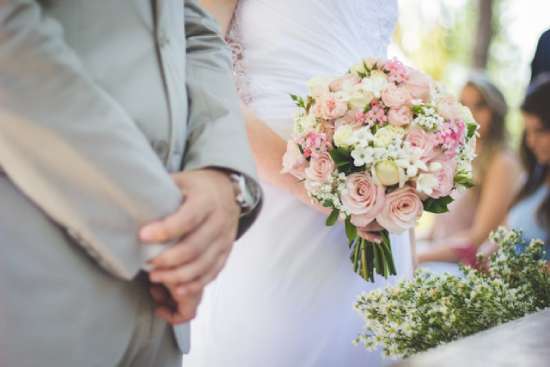 Bride and groom stand by one another at the altar, the bride holding a bouquet of pink and white roses.