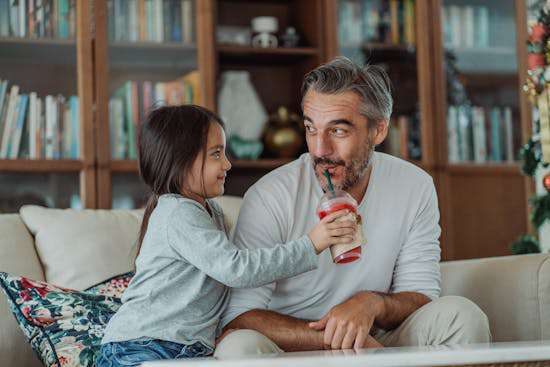 A smiling young girl holds a smoothie cup out to her father as he tastes it.