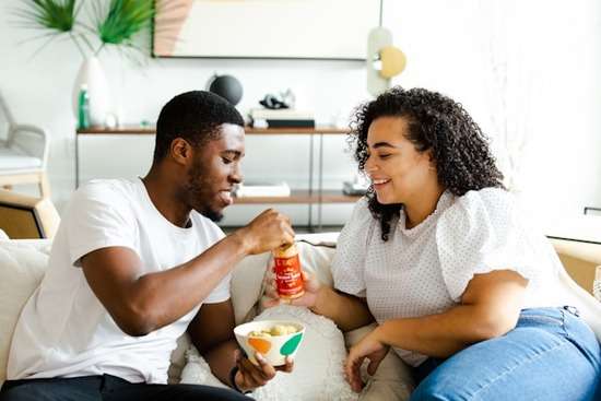 A cohabiting couple trade snacks as they sit together on their living room couch.