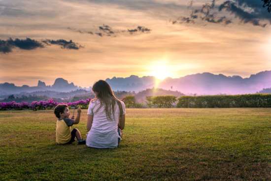 A mother sits in a field with her young son, attentively watching as he shows her something he found.