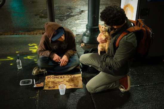 A man bending down, talking to a homeless person on the ground, and holding his cat, just like Jesus would