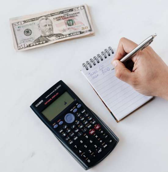 A person writes in a notebook beside a calculator and a stack of money as they evaluate their finances.