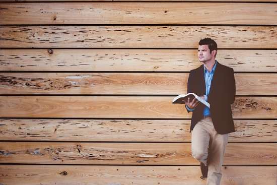 A man leaning against a wall reads his Bible, possibly thinking about what it says about God's appearance.